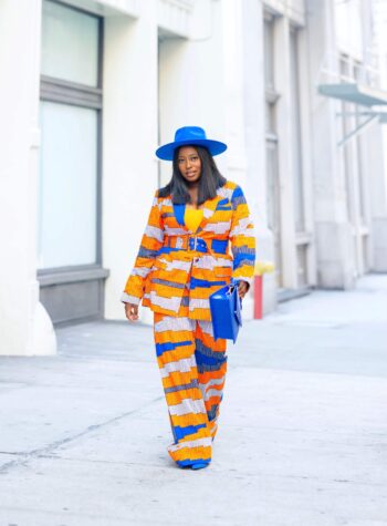 An woman in an orange and blue African print suit and cobalt blue hat stands on sidewalk